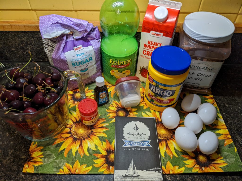 The ingredients for a pavlova: cherries, sugar, lemon, heavy cream, cocoa powder, corn starch, salt, almond extract, cream of tartar, eggs, and chocolate, all arrayed on a placemat.