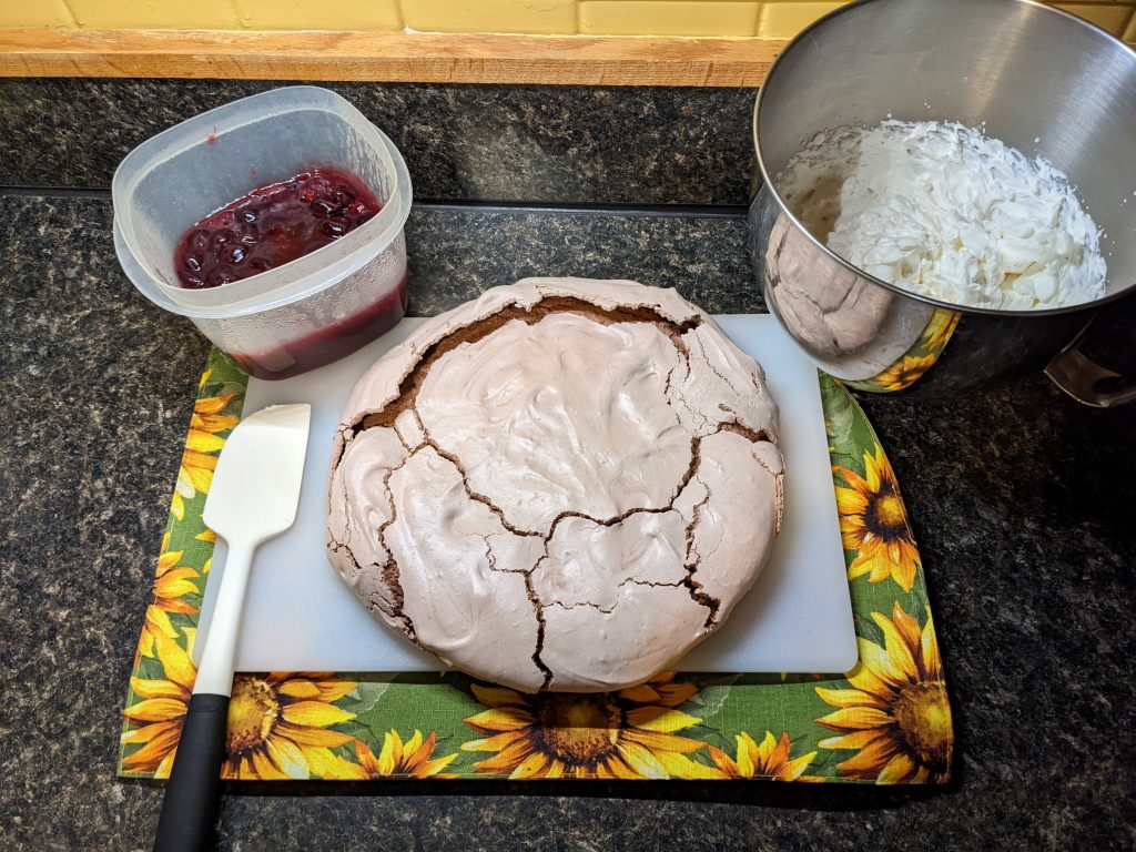 A baked pavlova with a bowl of whipped cream and a container of cherry sauce.
