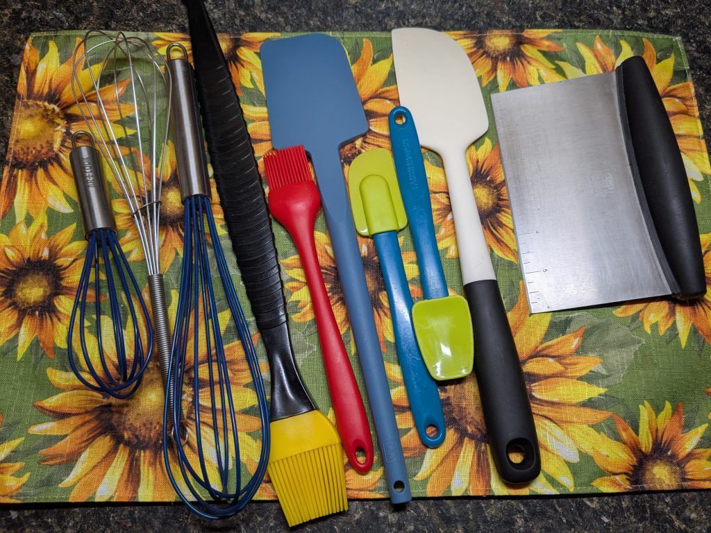 A photo of various baking utensils, including three whisks, two pastry brushes, four silicone spatulas, and a bench scraper.