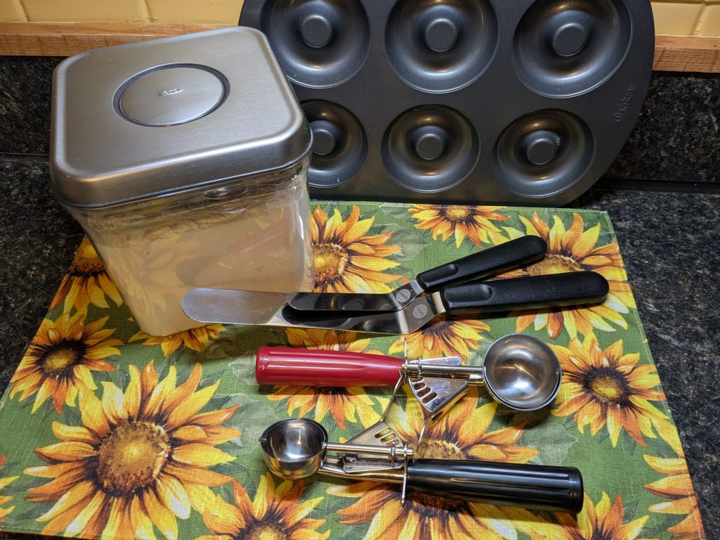 A photo of some optional baking accessories, including an ingredient keeper full of flour, a doughnut pan, icing spatulas, and cookie scoops.