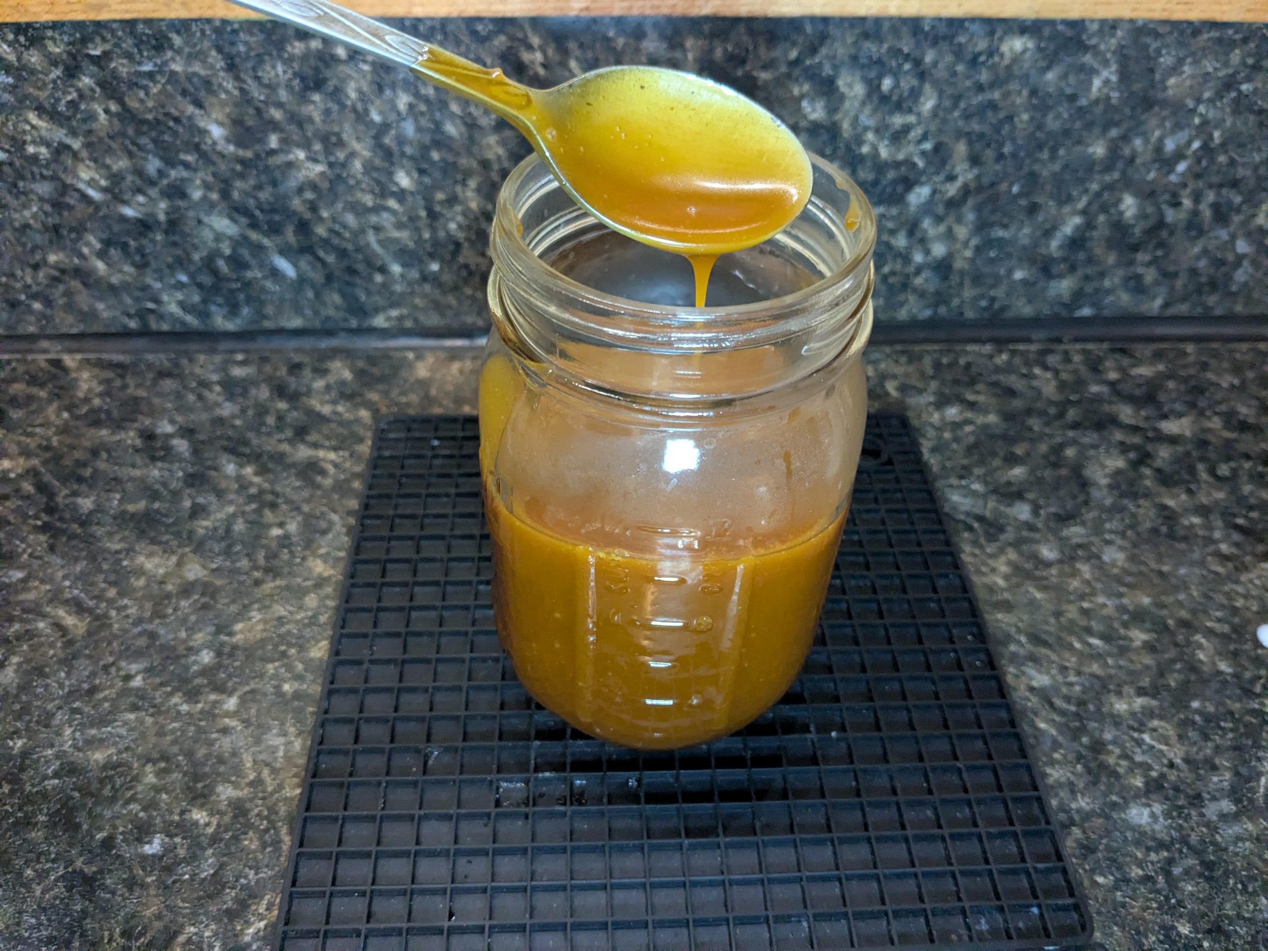 A half-full pint jar of salted caramel sauce, a spoonful streaming into the jar from above.