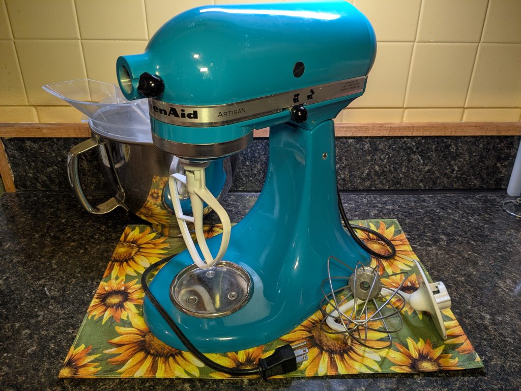 A photo of a teal Kitchenaid Artisan stand mixer with the basic attachments.