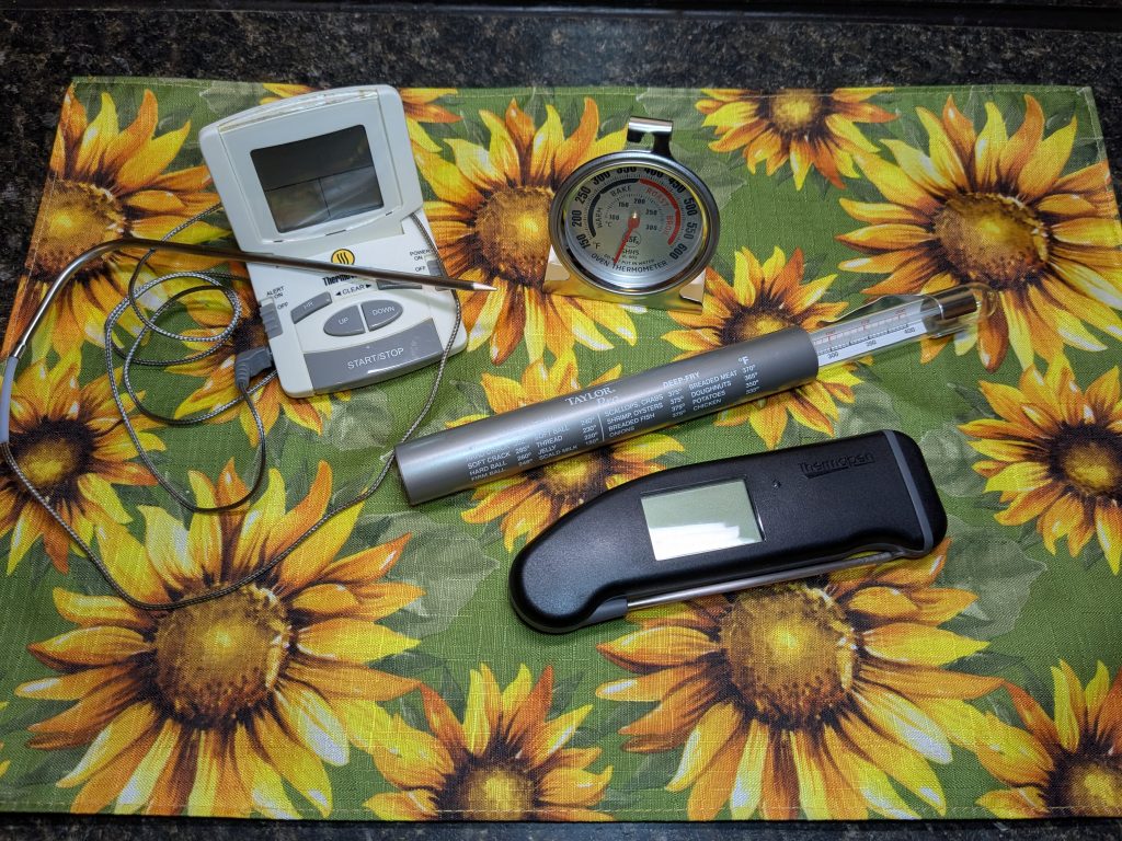 A photo of four thermometers; a digital probe thermometer, a Thermapen, an oven thermometer, and a candy thermometer.